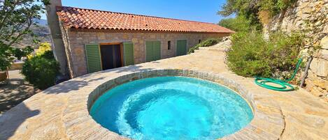 A stone pool with a tiled roof and green shutters, surrounded by stone walls and vegetation.