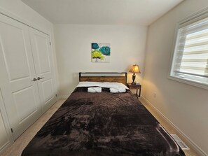 A bedroom with a large bed, a bedside table with a lamp, a painting on the wall, and a window with blinds.