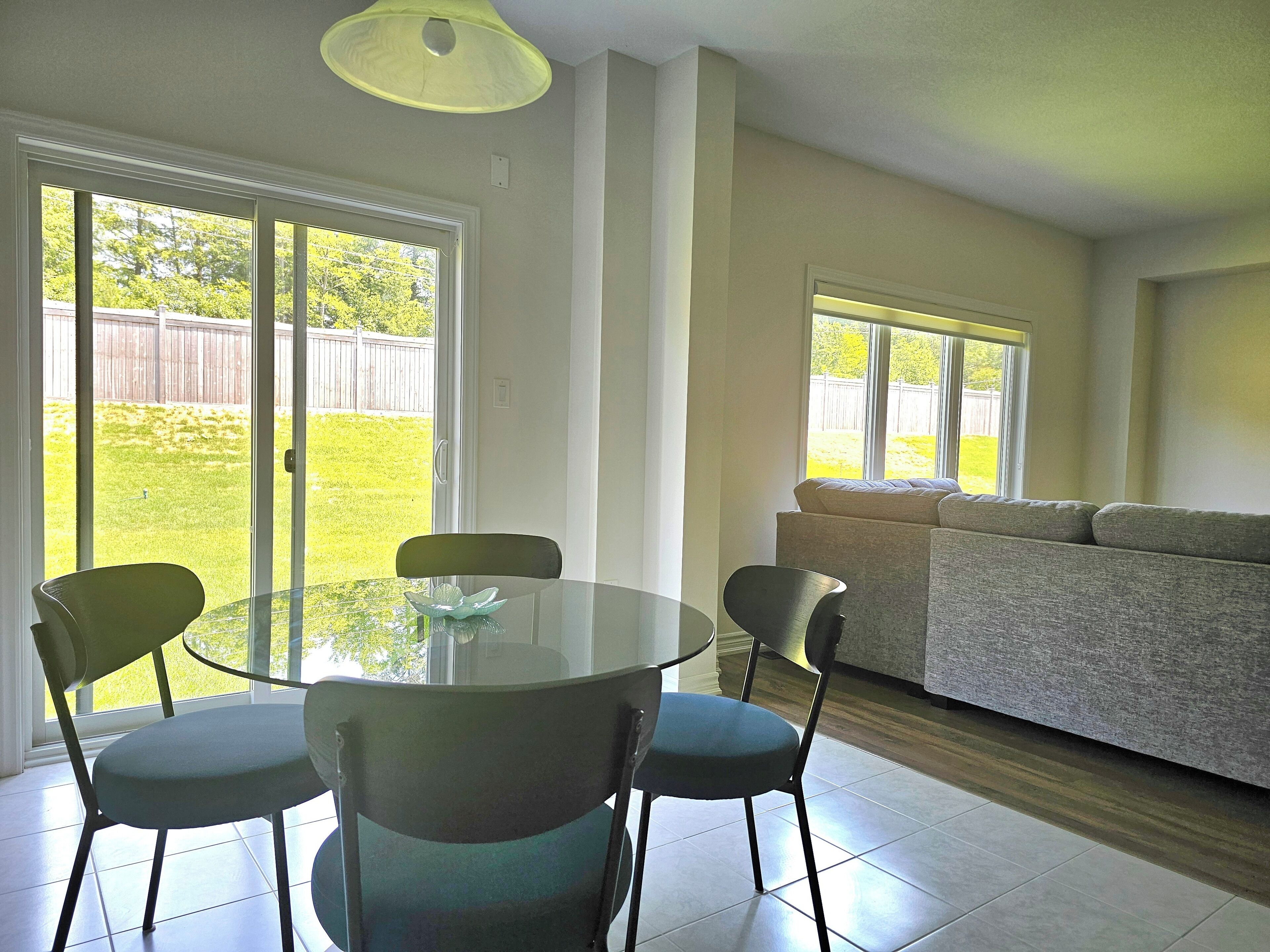 A dining area with a glass table and chairs, a sofa, and a view of a backyard through sliding glass doors.