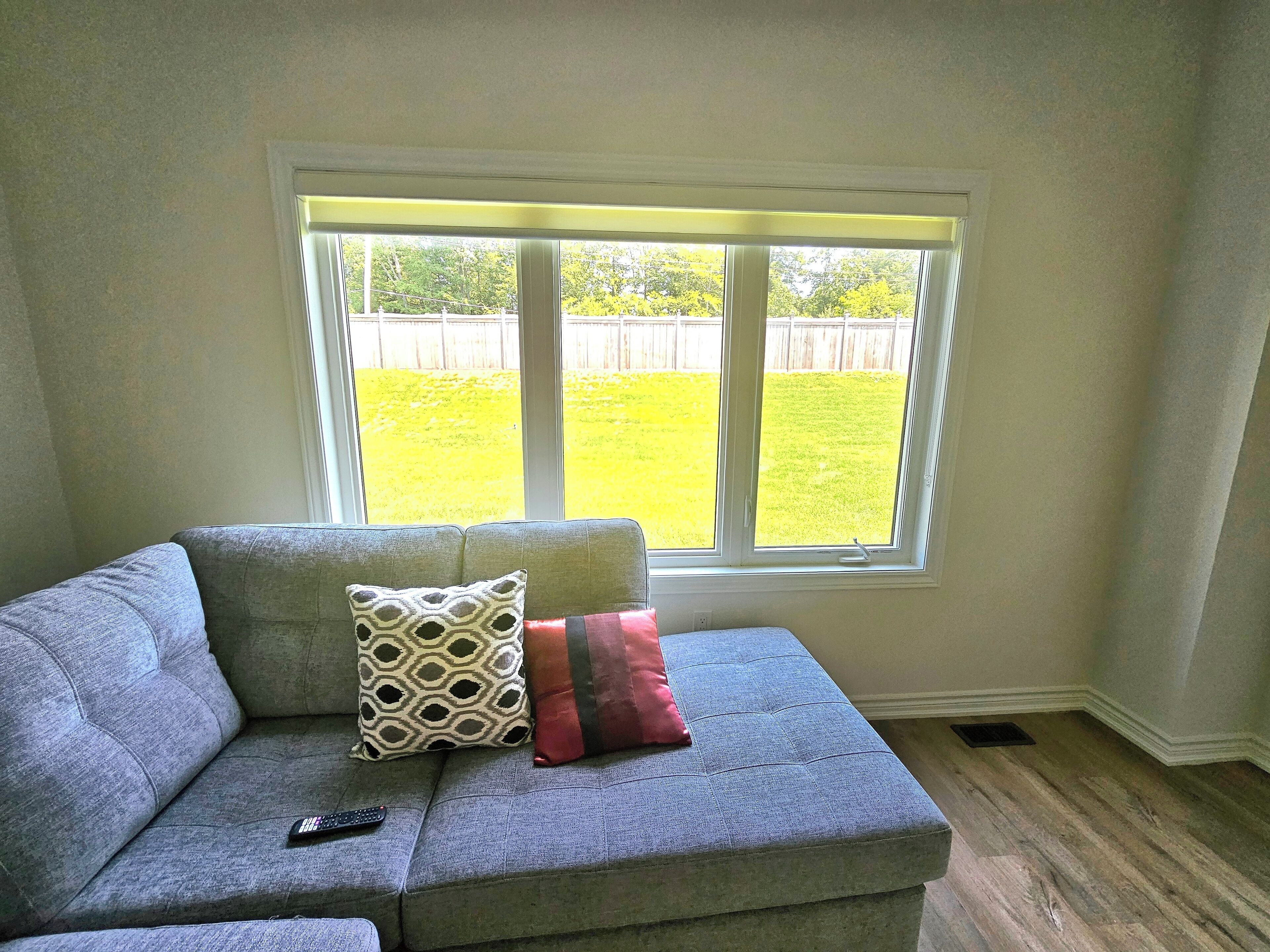A grey sectional sofa with patterned and solid cushions, a remote control on it, and a window with a view of a yard and wooden fence.