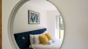 Une chambre avec un miroir rond, un lit avec une literie blanche et jaune, une tête de lit bleue et un tableau abstrait encadré accroché au mur.