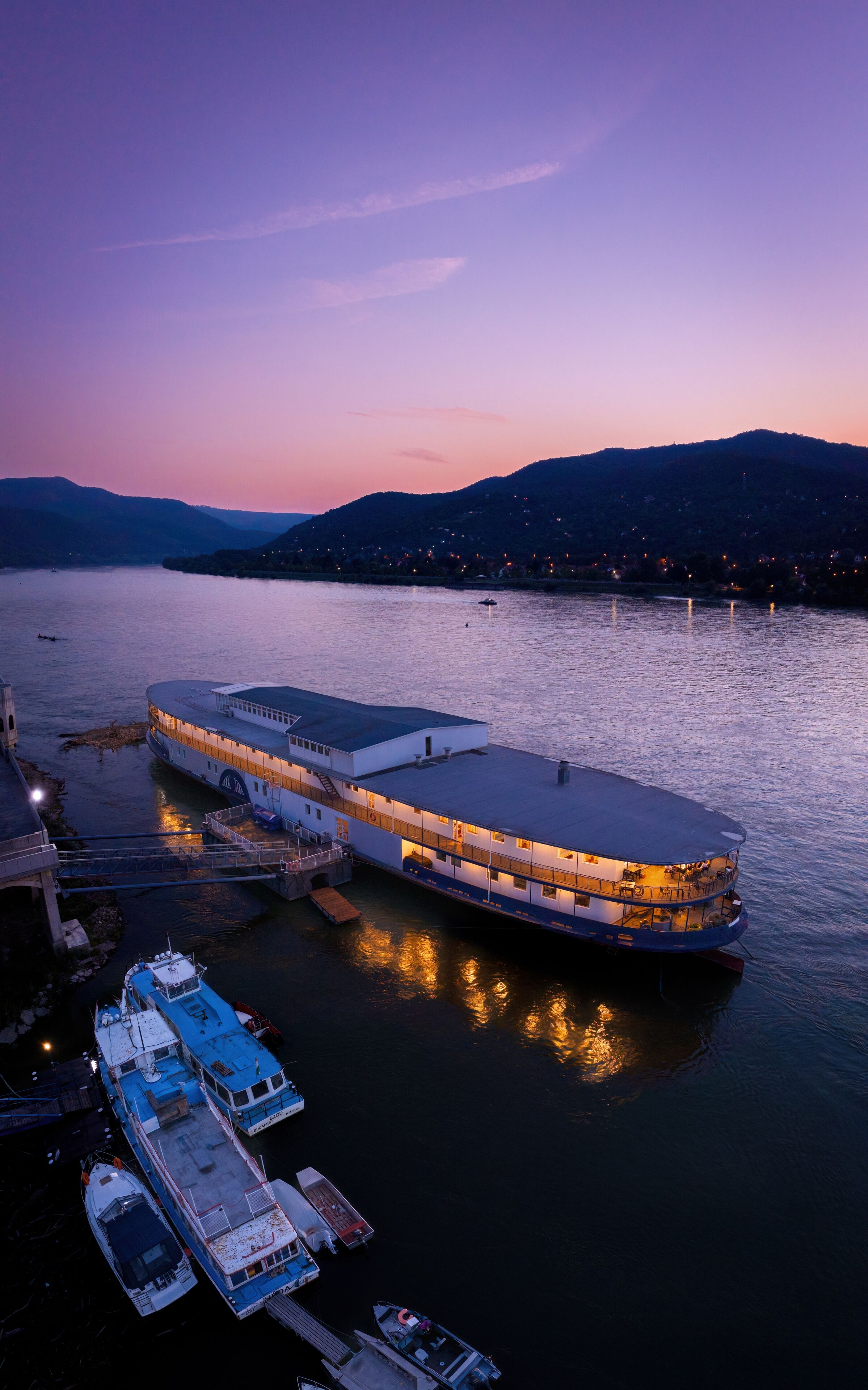 A large riverboat docked at a pier with multiple levels of accommodation, surrounded by water and distant hills.
