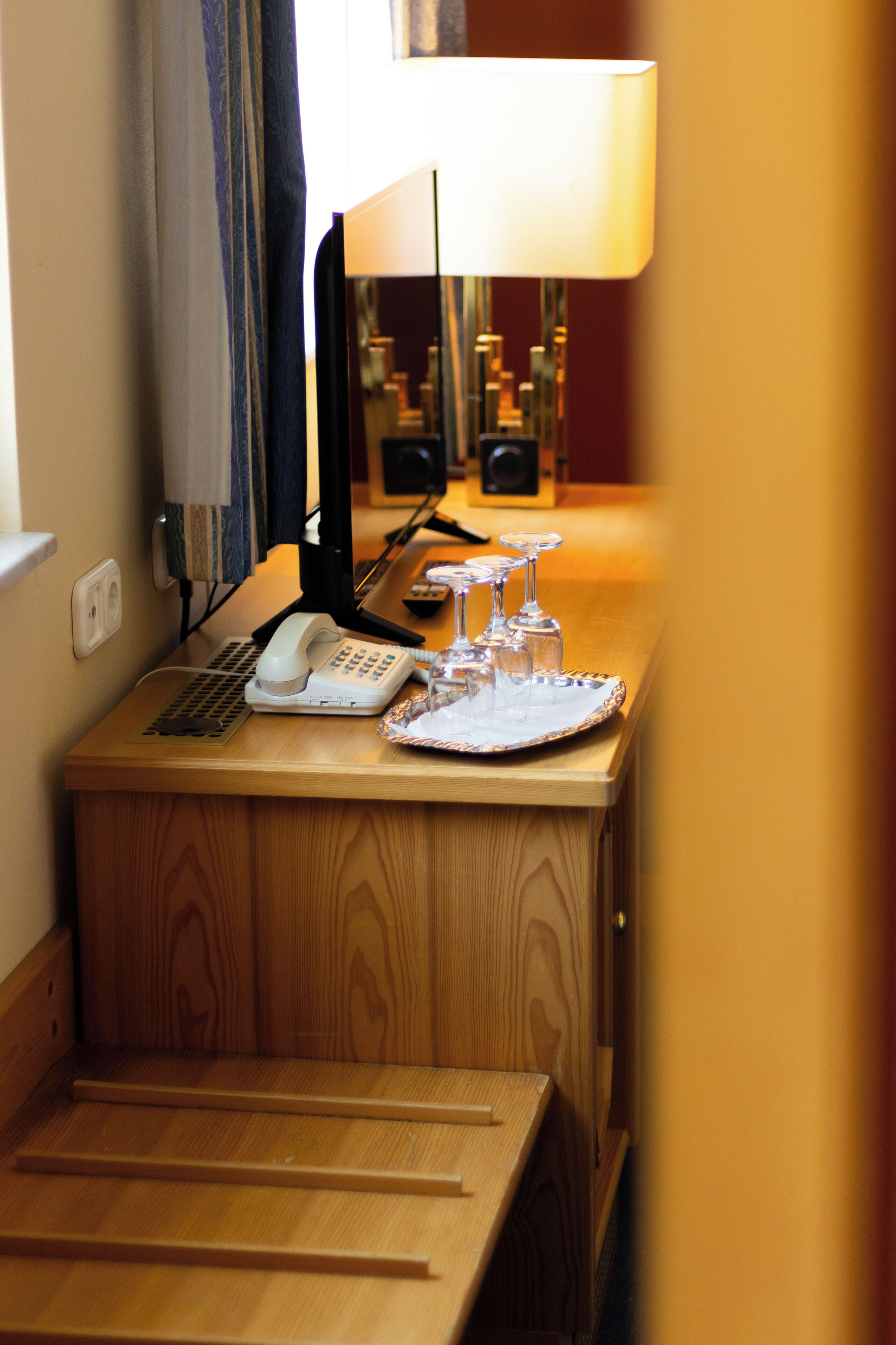 A wooden desk with a telephone, a lamp, and a tray with wine glasses.