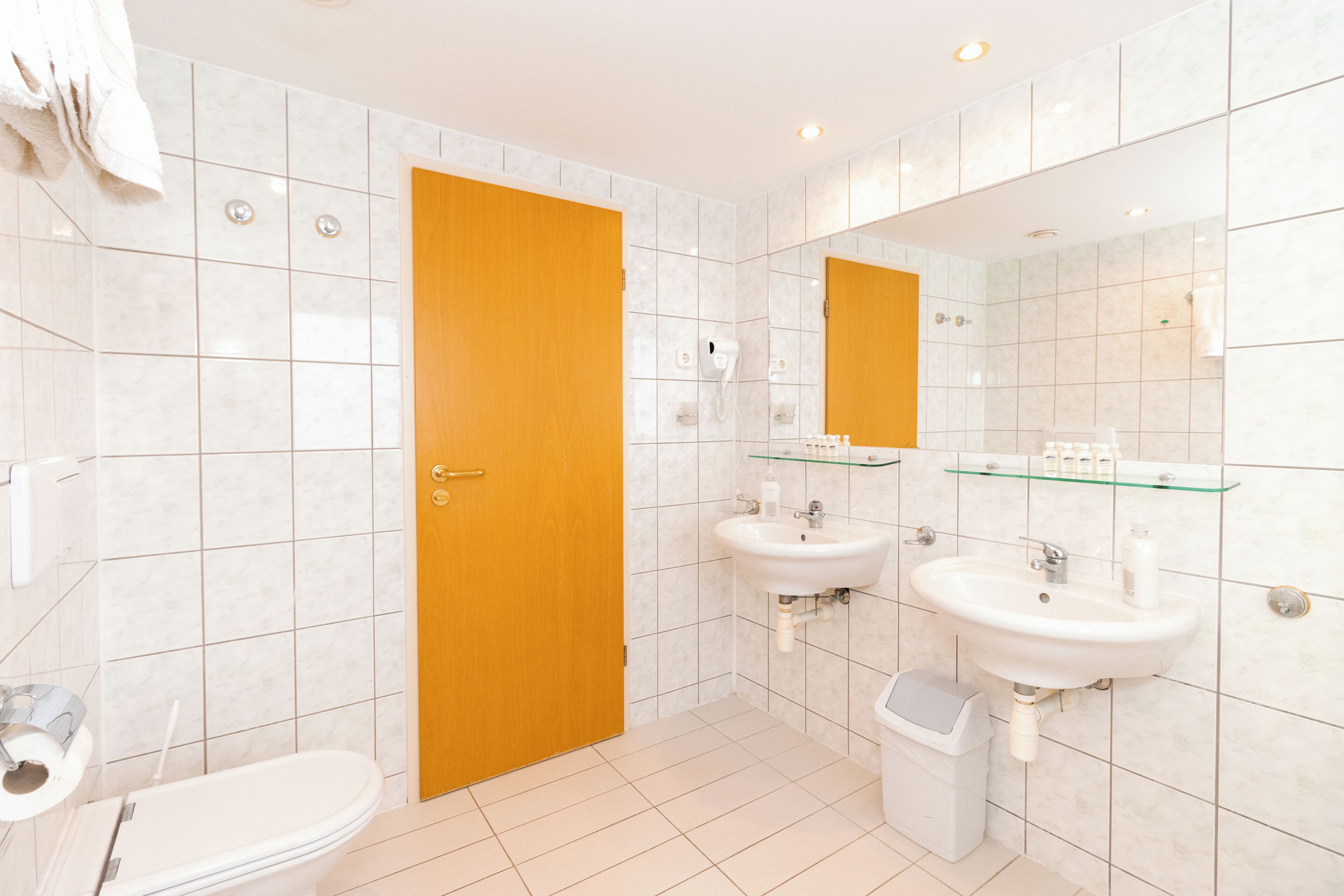 A bathroom with a toilet, two sinks, a glass shelf, and a wooden door.