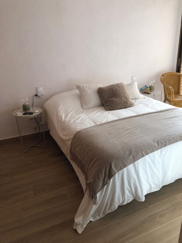 Un lit bien fait, recouvert d’une couverture blanche et grise, une petite table de chevet avec une lampe et un fauteuil en osier.