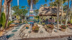 A tropical resort with a thatched-roof hut, palm trees, and a signboard with various amenities.