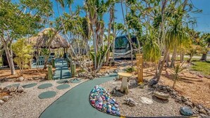 A tropical resort with a thatched-roof hut, palm trees, a paved walkway, and a bus in the background.