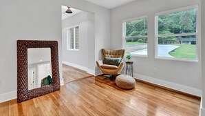 A spacious room with hardwood flooring, a large mirror, a tufted armchair, and a view of a green landscape through large windows. - Modern Farmhouse in the City -  4-bedroom house in charming Atlanta (Atlanta)