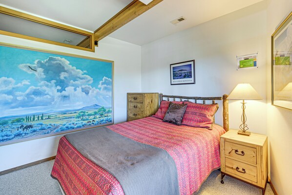Un dormitorio con un cuadro de paisaje grande, una cama con colcha roja y rosa, una mesita de noche de madera con una lámpara, y un cuadro enmarcado en la pared.