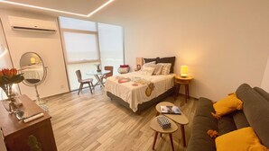 A modern hotel room with a bed, a sofa, a small table, a chair, and a desk with a mirror and vase. - Acogedor estudio en Adamant Cabo Norte en Merida, perfecto parejas o home office (Mérida)