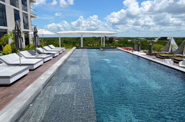 A swimming pool with lounge chairs and umbrellas, surrounded by greenery and a modern building.