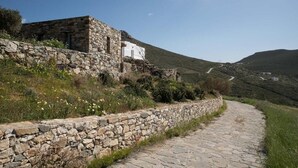 A stone pathway leading to a rustic stone building with a white door, surrounded by greenery and hills in the background.