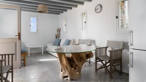 A modern living room with a glass-top coffee table, wooden chairs, a sofa with cushions, a hanging light fixture, and a clock on the wall.