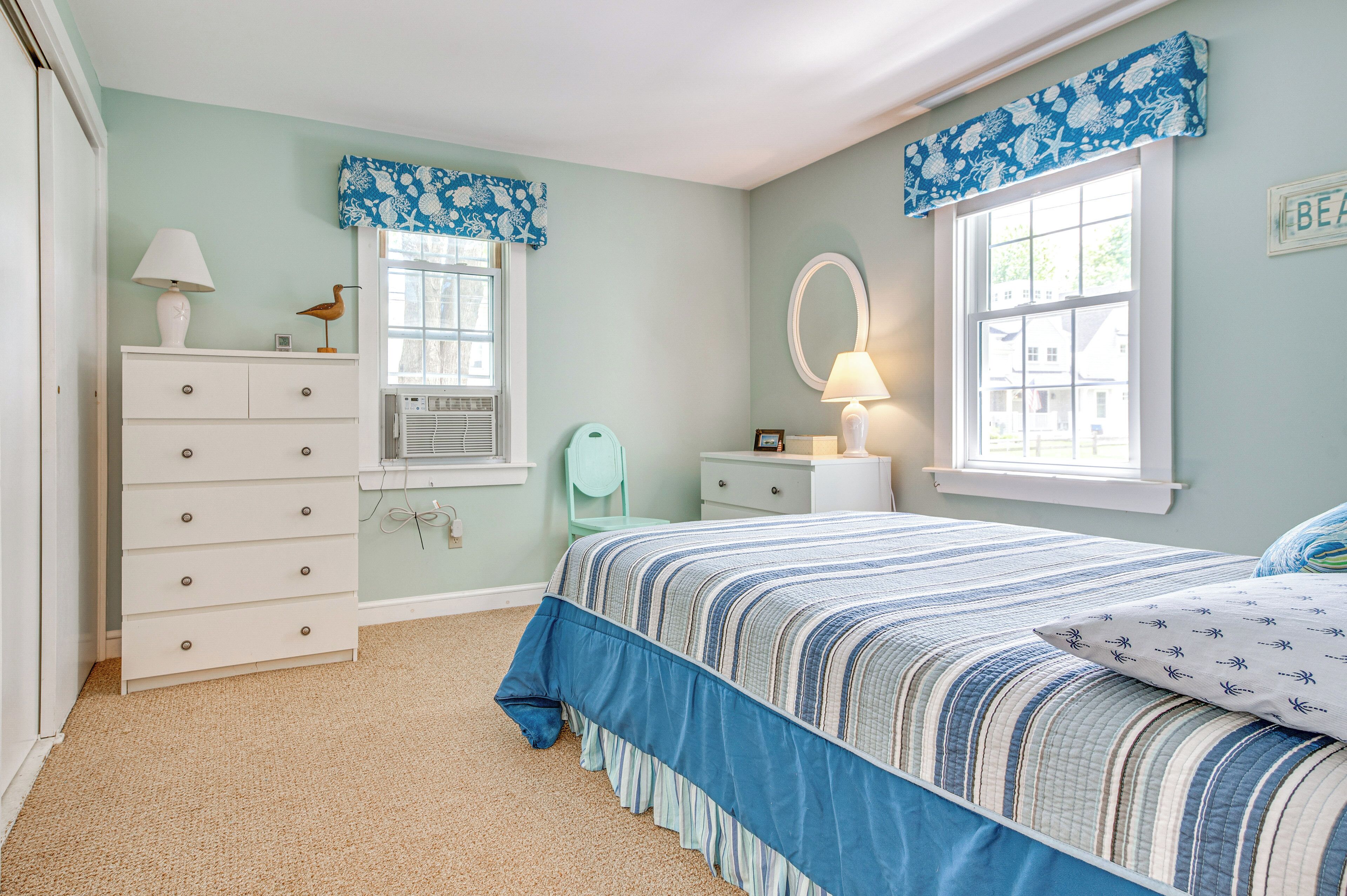 A bedroom with a bed, dresser, vanity, and two windows with curtains.
