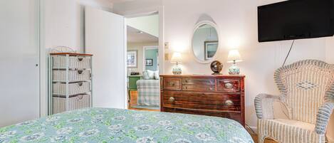 Une chambre à coucher avec une couvre-lit à motifs floraux, une commode en bois, un fauteuil à bascule et un téléviseur à écran plat fixé au mur.