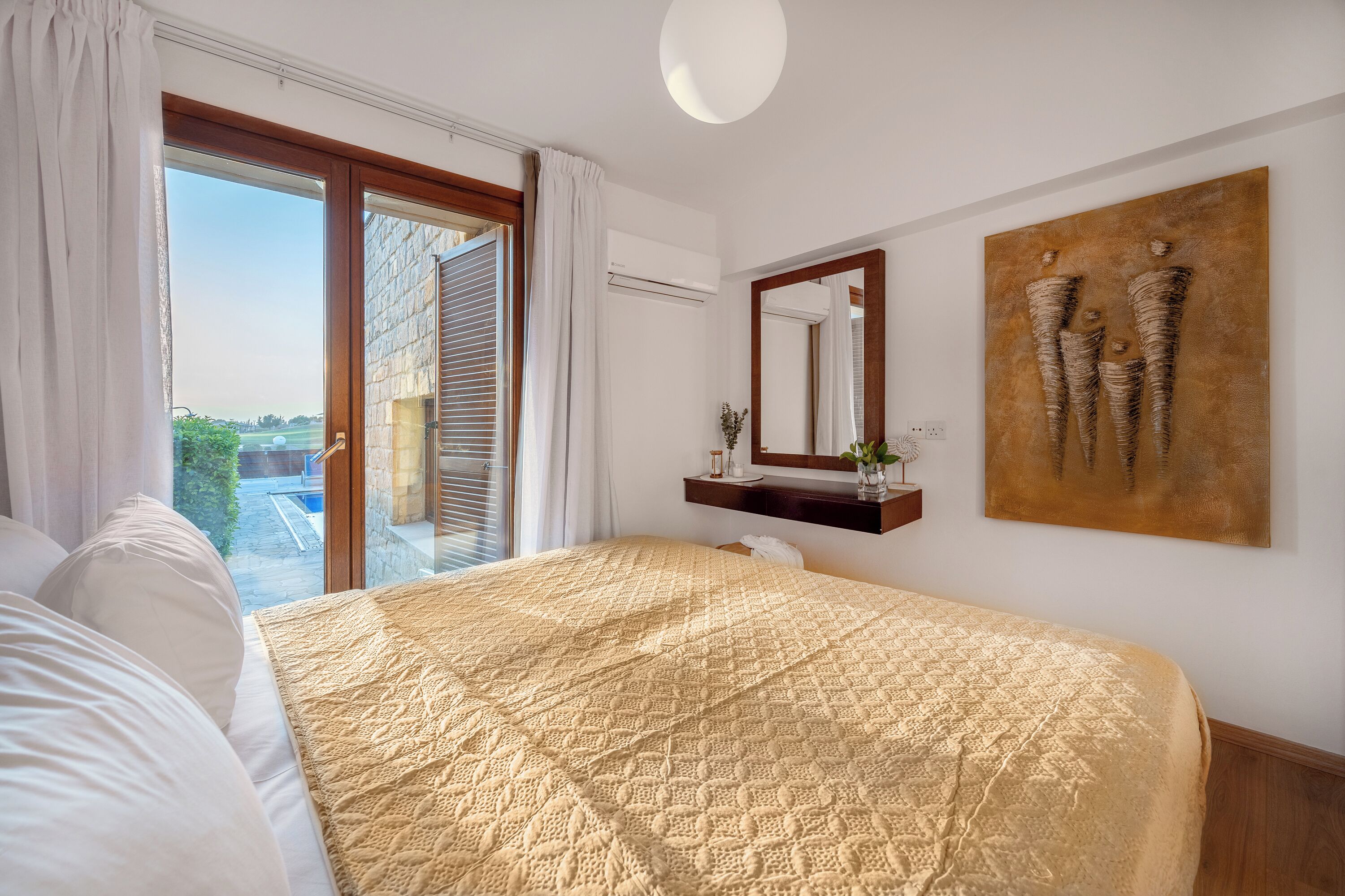 A bedroom with a large bed, a sliding glass door to a balcony, a wall-mounted mirror, and a piece of abstract art.