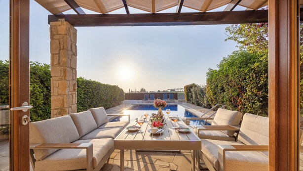 An outdoor patio with a stone pillar, cushioned seating, a table set for dining, and a view of a swimming pool. - Hg06 Adjacent to Aphrodite Hills- Private Pool (Kouklia)