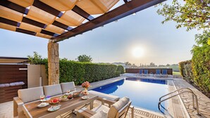 An outdoor pool area with a wooden deck, seating, and a stone wall. - Hg06 Adjacent to Aphrodite Hills- Private Pool (Kouklia)