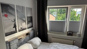 Une chambre à coucher avec un lit, une fenêtre avec des stores, un radiateur et des œuvres d’art accrochées au mur.