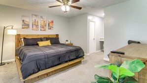 Un dormitorio con cama, ventilador de techo, mesita de noche, lámpara, planta y cómoda de madera.