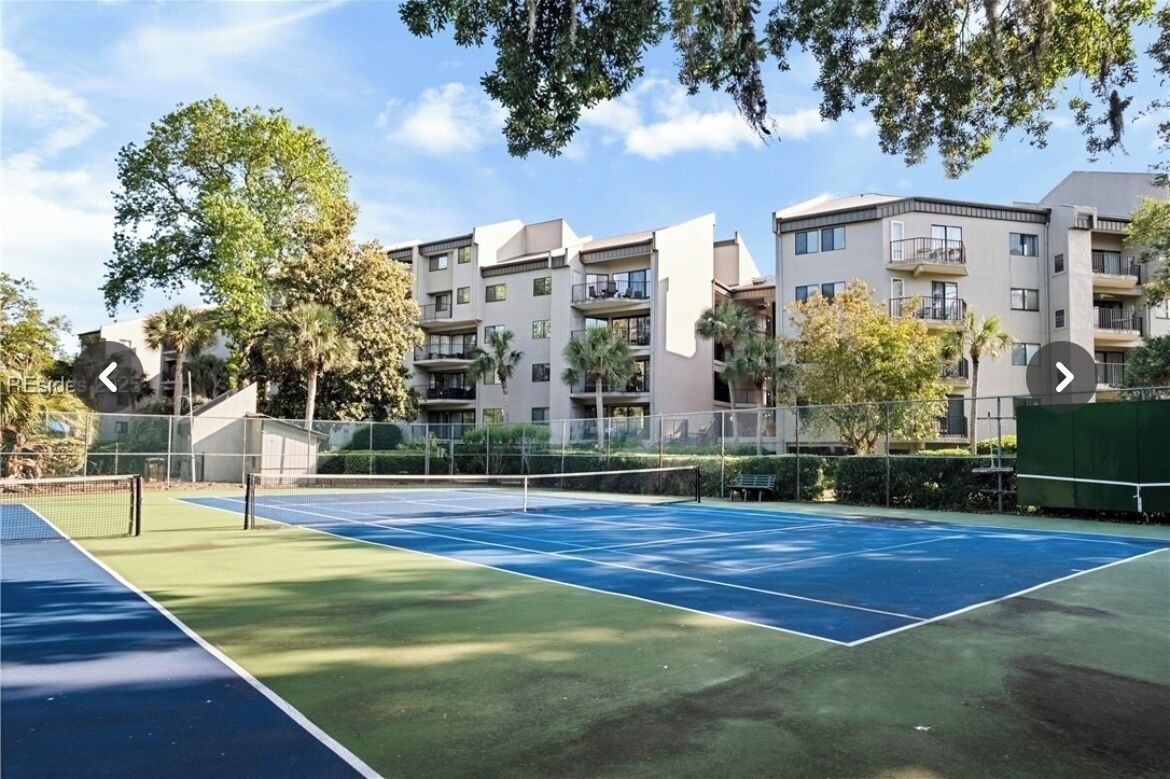 Una cancha de tenis con red, rodeada de árboles y un edificio de varios pisos con balcones.