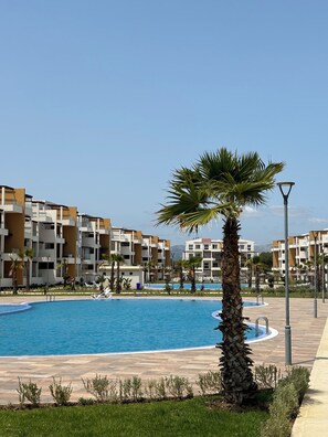 Ein Swimmingpool, umgeben von modernen Mehrfamilienhäusern, mit einer Palme und einer Laterne im Vordergrund.