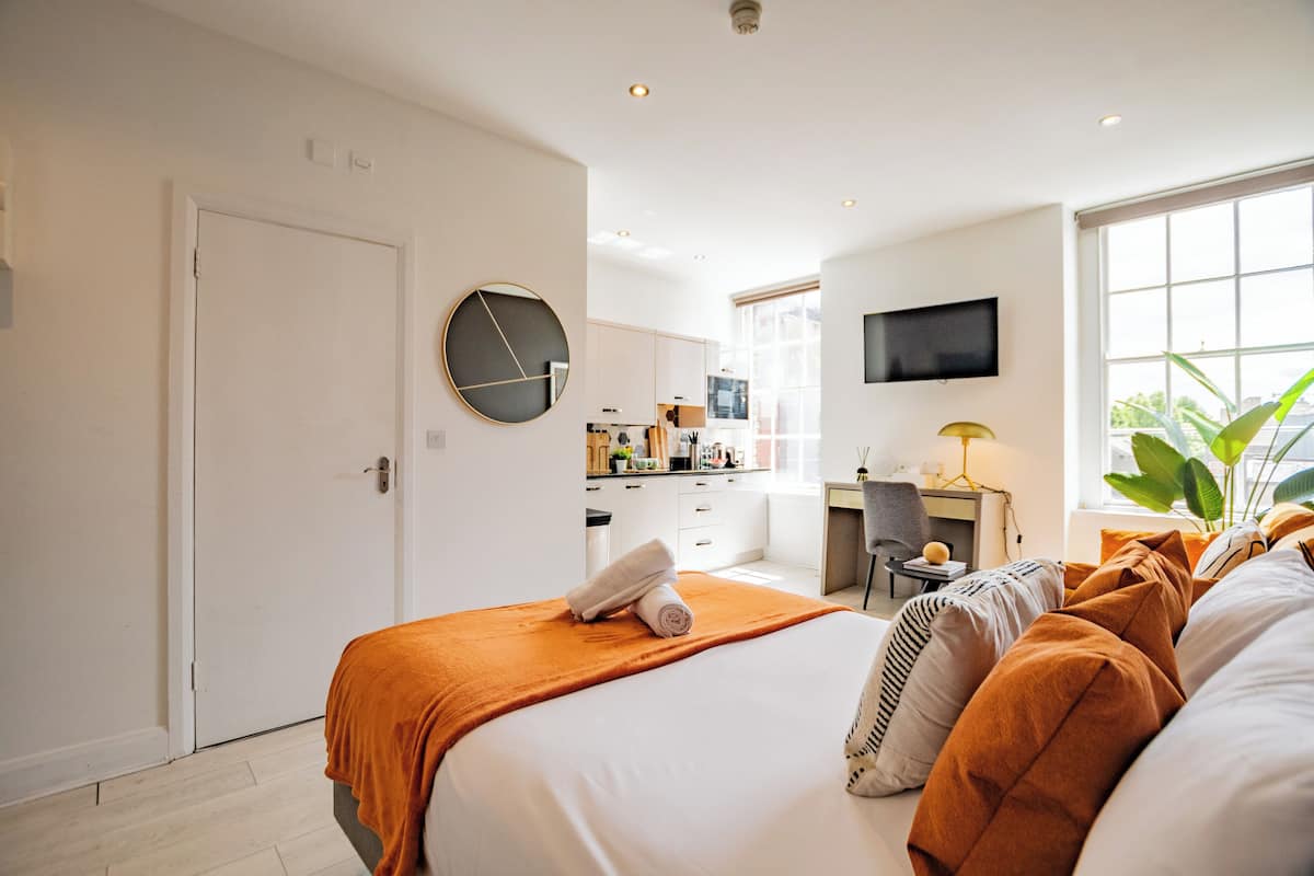 A modern hotel room with a bed, a desk, a television, and a view of the kitchen.