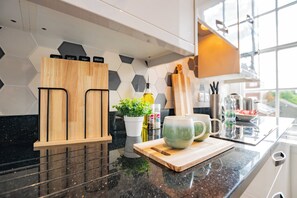 Eine Küche mit schwarzer Granitabdeckung, Schneidebrett aus Holz, einer grünen Tasse, einem Topfpflanzen und einer Rückwand mit sechseckigen Fliesen.