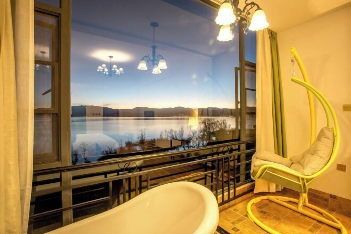 A bathroom with a bathtub, a hanging chair, and a view of a lake and mountains.