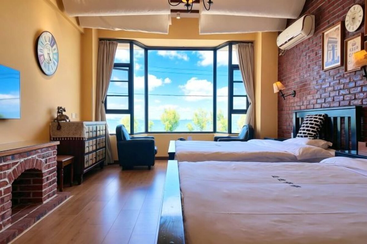 Una habitación con chimenea de ladrillo, dos camas, un ventanal con vistas y un reloj en la pared.