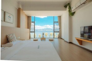 A modern hotel room with a large bed, a TV, and a view of the city through large windows.