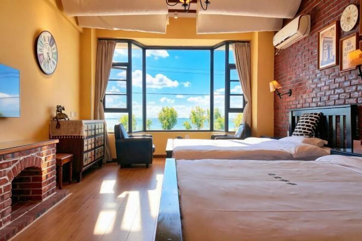 Une pièce avec une cheminée en briques, deux lits, une fenêtre donnant sur un beau paysage et une horloge au mur.