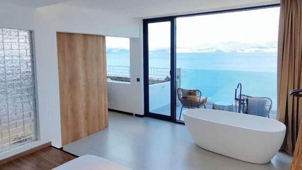 Um banheiro moderno com uma banheira de pé, uma janela ampla com vista para o mar e uma porta de vidro deslizante que dá para uma varanda com mobiliário externo.