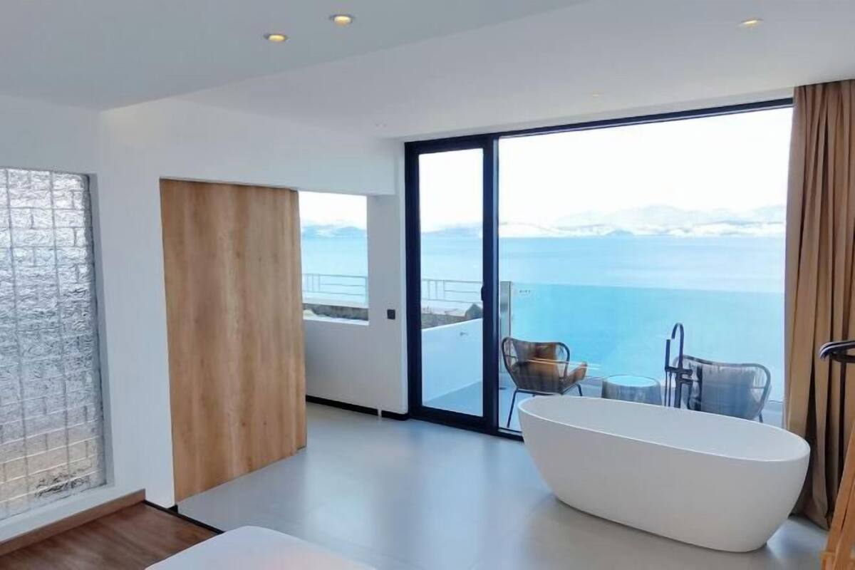 Une salle de bain moderne avec une baignoire sur pieds, une grande fenêtre donnant sur la mer et une porte-fenêtre coulissante qui mène à un balcon aménagé avec des sièges d’extérieur.