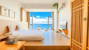 A modern hotel room with a large bed, a desk, a TV, and a view of the sea.