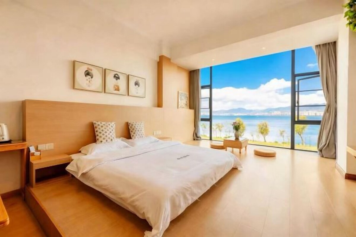 Una habitación de hotel moderna con una cama grande, una mesita de noche, un ventanal con vista al mar y una pequeña zona de estar.