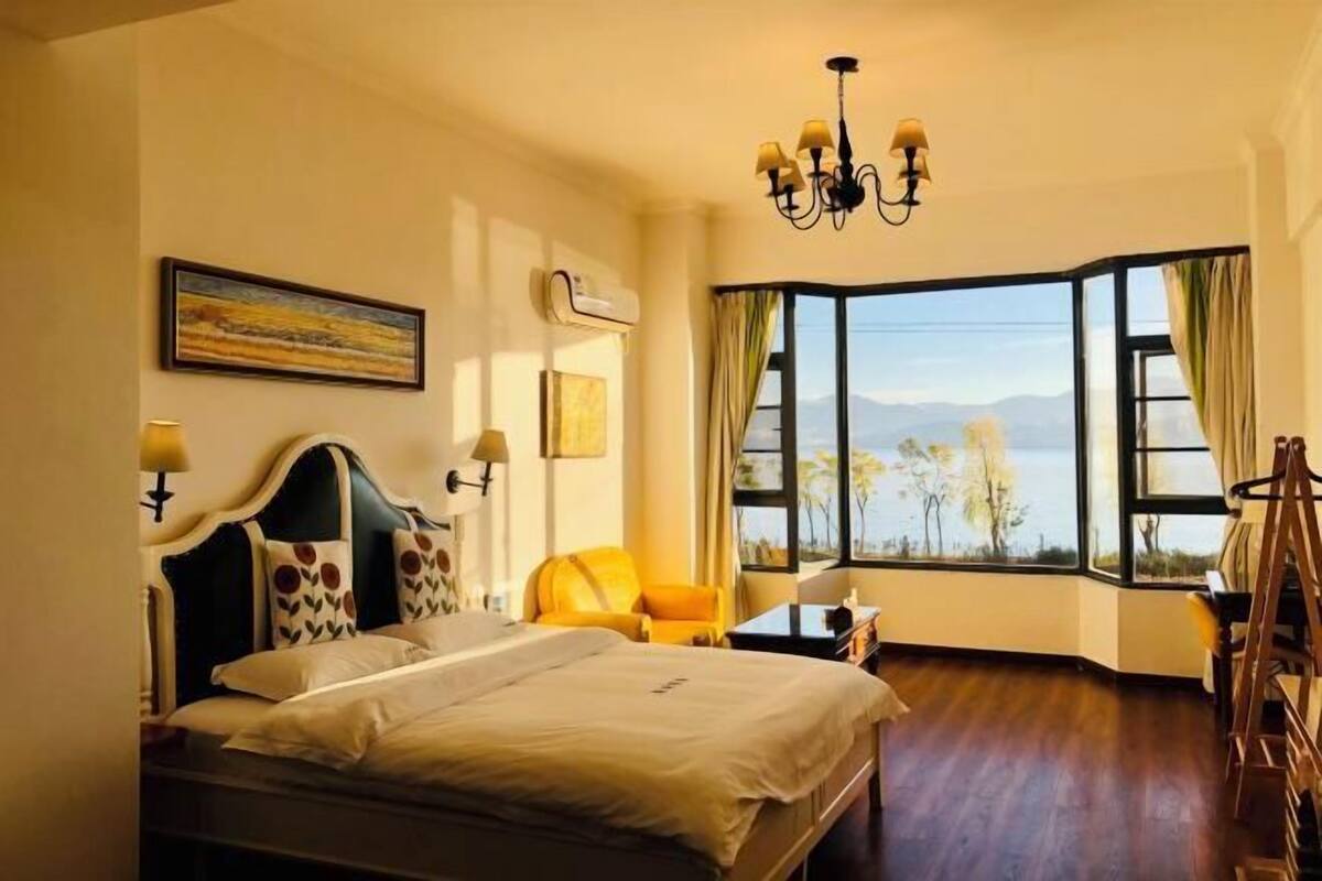 Une chambre d’hôtel comprenant un lit, un bureau, une chaise, un lustre, une grande fenêtre avec vue et des œuvres d’art accrochées au mur.