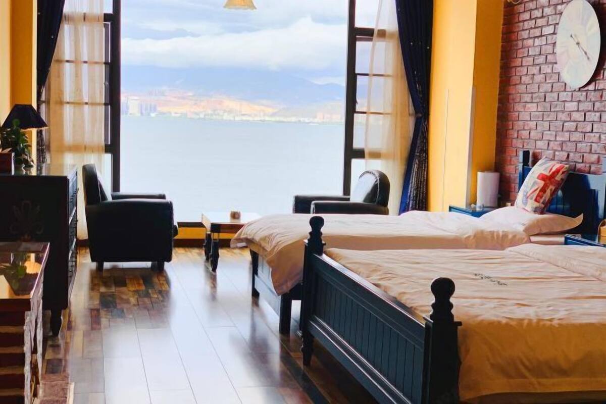 Une pièce dotée d’une grande fenêtre donnant sur une étendue d’eau, avec un lit, une chaise, une petite table et une lampe.