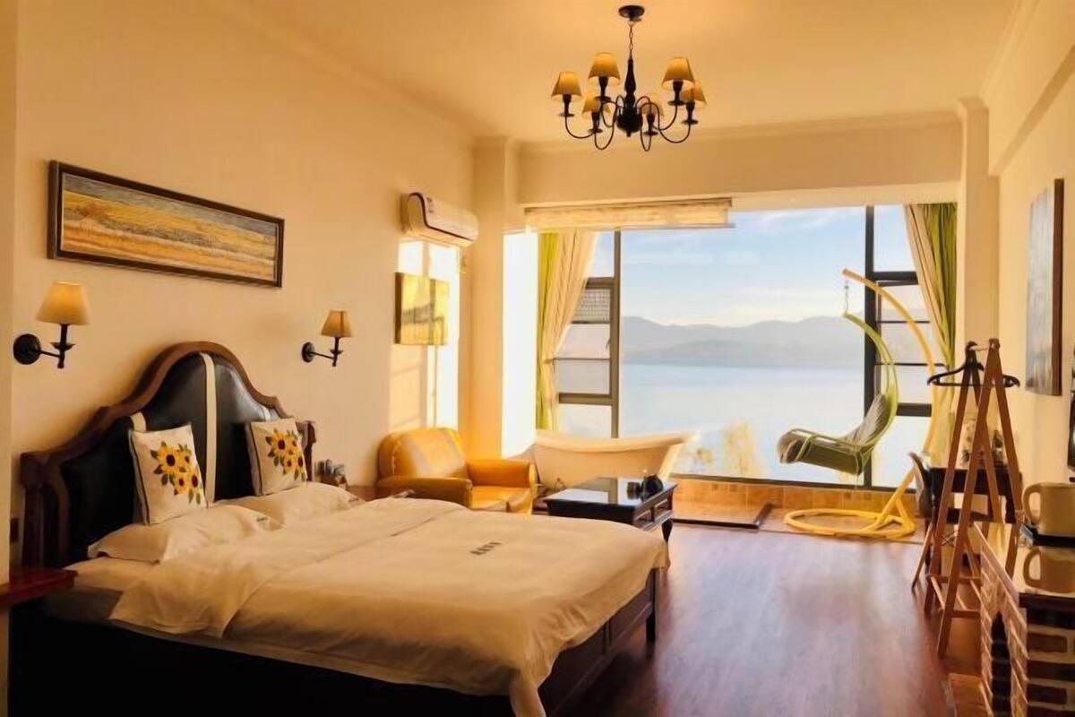 Une chambre d’hôtel avec un grand lit, un bureau, une chaise, une fenêtre donnant sur la mer et un lustre.