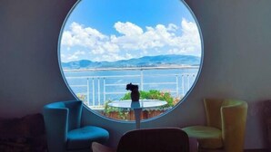 Quarto com janela redonda que oferece vista para um corpo de água, montanhas e uma ponte. Há duas cadeiras e uma pequena mesa com um vaso.