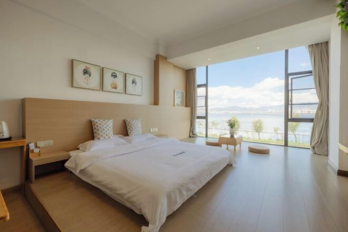 A modern hotel room with a large bed, a bedside table, a nightstand, a view of the outdoors, and a window with curtains.