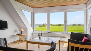 Een kamer met een schuine plafond, grote ramen met uitzicht op een groen veld, een tafel met een kaars en een televisie aan de muur.