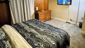 A bedroom with a bed, a wooden dresser, a television mounted on the wall, and a floor lamp. - Townhome with Game Zone, Big Beds, & Smart TVs (Westmont)