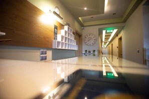 A modern hotel lobby with a wooden reception desk, a clock on the wall, and a long corridor with doors. - Amalia Center (Volvi)