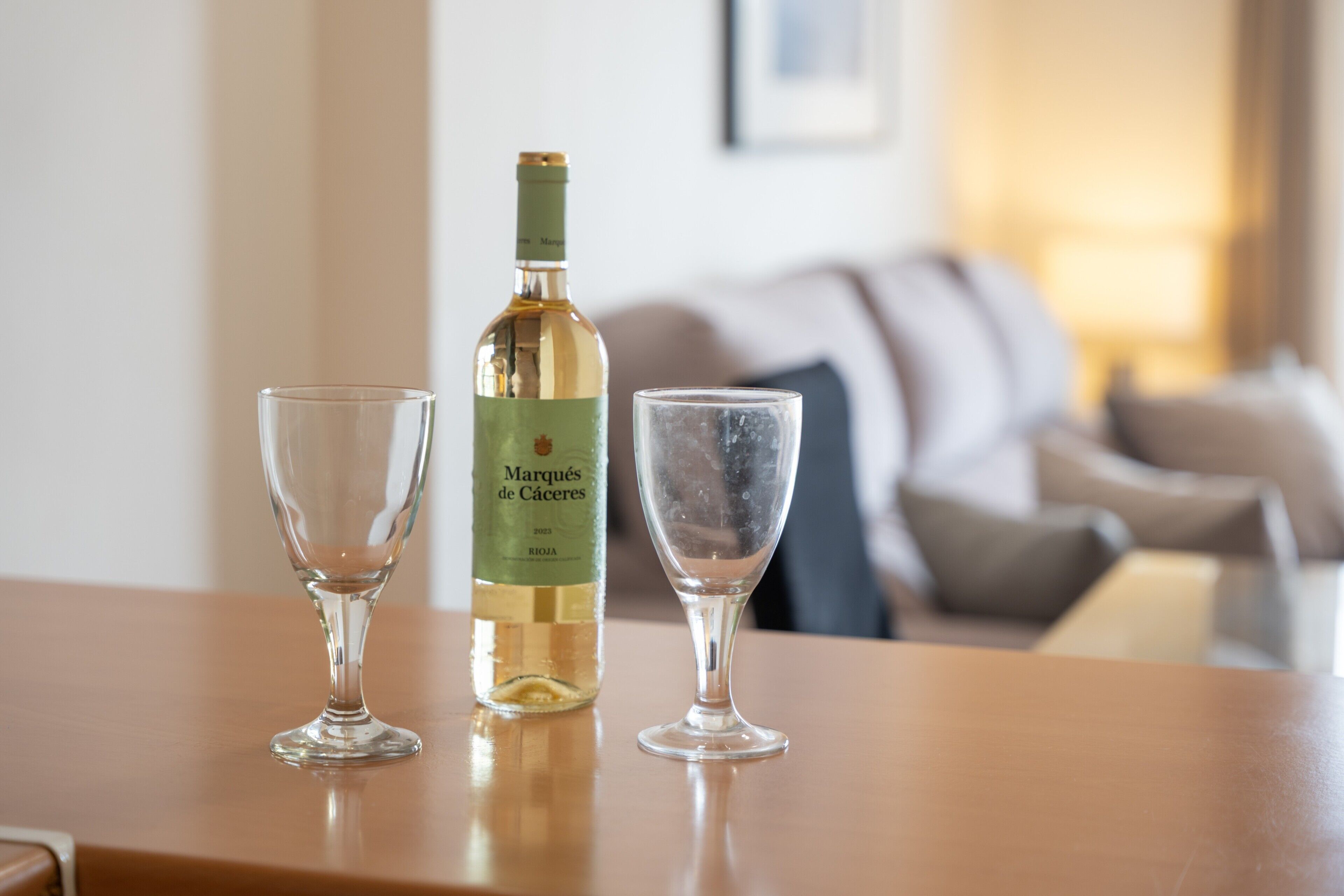 A bottle of Marqués de Cáceres wine and two glasses on a wooden table in a living room.