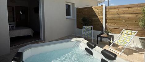 Un bain à remous dans un jardin, avec une clôture en bois, une chaise longue et une petite table agrémentée d’une plante.