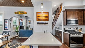 A modern kitchen with a central island, wooden cabinets, stainless steel appliances, and a dining area with a blue sofa and wooden table. - Private Pool & Hot Tub • 2 King Suites • No Cleaning Fee • Pet Friendly (Galveston)