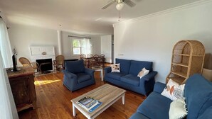 Un salon avec deux canapés bleus, une table basse, des fauteuils en osier, une cheminée et un coin repas avec une table et des chaises.
