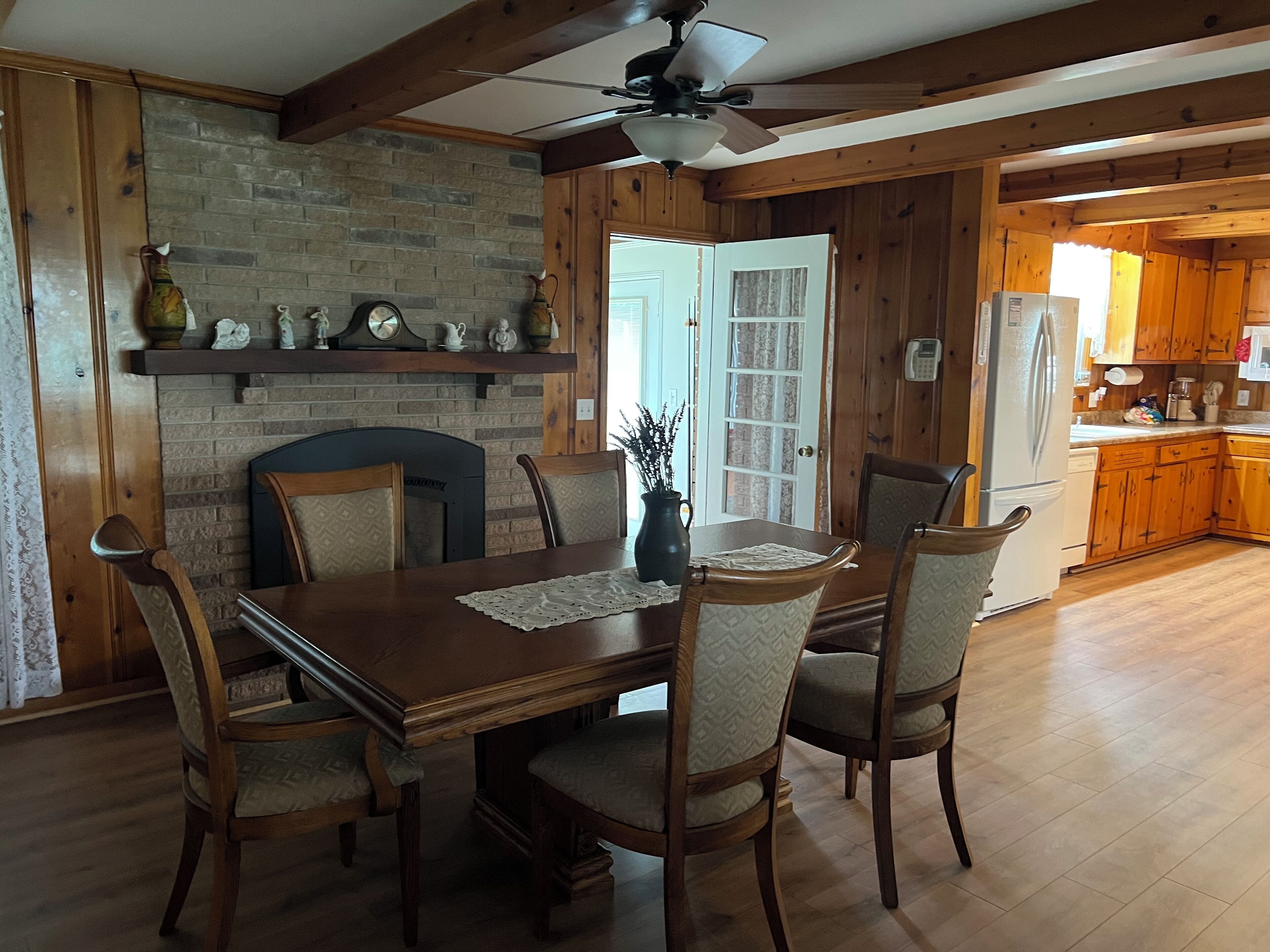 Uma área de refeições com mesa e cadeiras de madeira, lareira de tijolos e ventilador de teto.