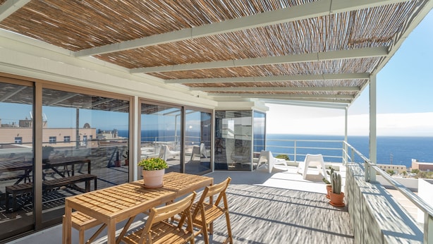 A wooden deck with a table and chairs, potted plants, and a view of the sea.